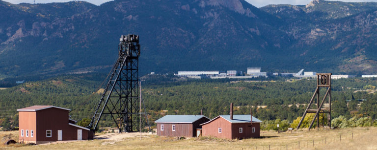 Old mining structures with a wooden elevator and small red buildings in a grassy field, backed by forested hills and distant industrial facilities.