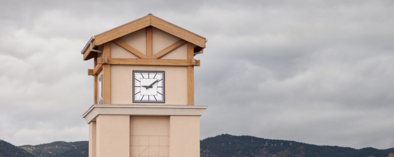 A beige clock tower with wooden beams stands against a cloudy sky and distant mountains.