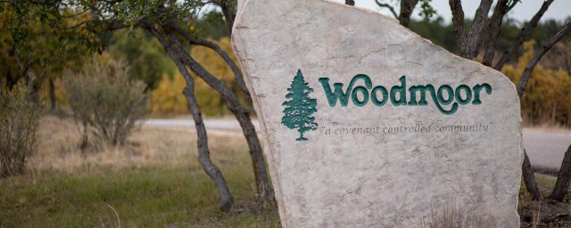 Sign with the text "Woodmoor: a covenant controlled community" on a large rock, surrounded by trees and grass.